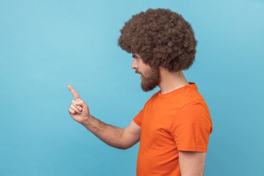Side view of serious man with Afro hairstyle wearing orange T-shirt standing with admonishing gesture, scolding for mistake and warning. Indoor studio shot isolated on blue background.