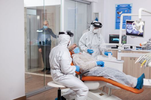 Senior patient wearing hazmat suit during coronavirus in dentist office Elderly woman in protective uniform during medical examination in dental clinic.