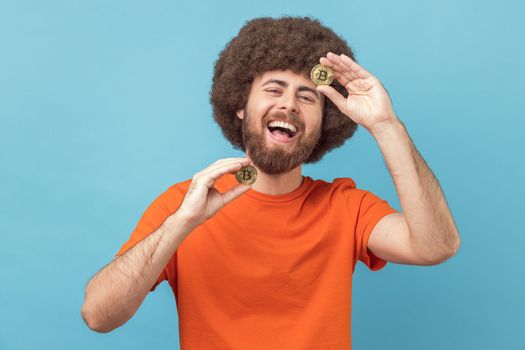 Portrait of funny laughing man with Afro hairstyle wearing orange T-shirt standing holding bitcoins, paying attention at new digital cryptocurrency. Indoor studio shot isolated on blue background.