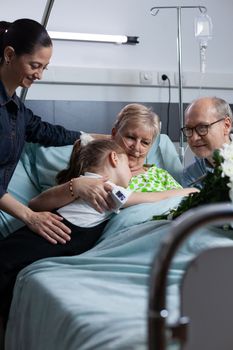 Elderly woman suffering illness lying in hospital observation room bed, hugging young granddaughter during relatives visit. Family giving flowers bouquet to sick grandmother at medical clinic.