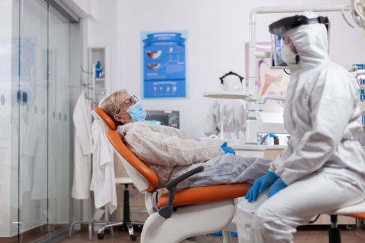 Dentist talking about teeth hygiene wearing hazmat suit during cornavirus outbreak. Elderly woman in protective uniform during medical examination in dental clinic