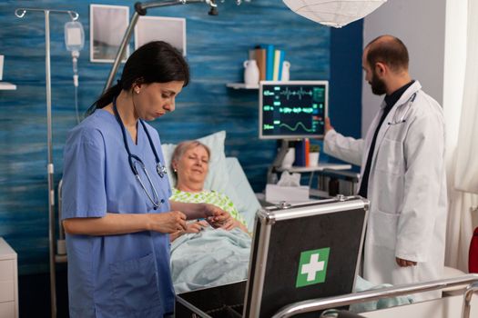 Female nurse preparing medicine with syringe to apply to sick elderly woman in hospital room bed. Male doctor analyzing vital sign values displayed on monitor, professional health care.