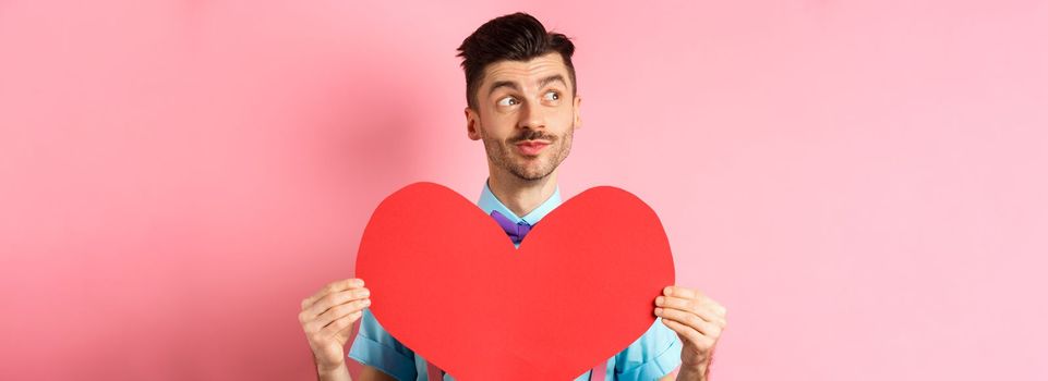 Valentines day concept. Dreamy romantic man looking left and showing big red heart cutout, standing on pink background.