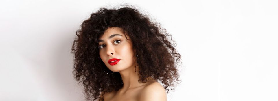 Close-up of beautiful caucasian woman with curly haircut and red lipstick, looking tender at camera, standing over white background.