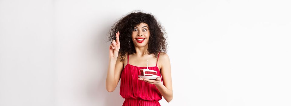 Excited birthday girl in red dress, cross fingers while making wish and blowing candle on bday cake, smiling happy, white background.