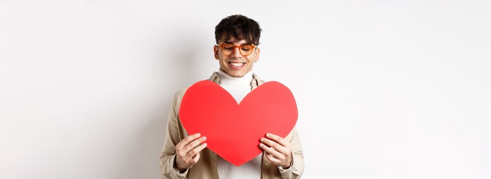 Handsome and stylish man in sunglasses, prepare surprise postcard for girlfriend on Valentines day, holding big red heart cutout and smiling happy, standing over white background.