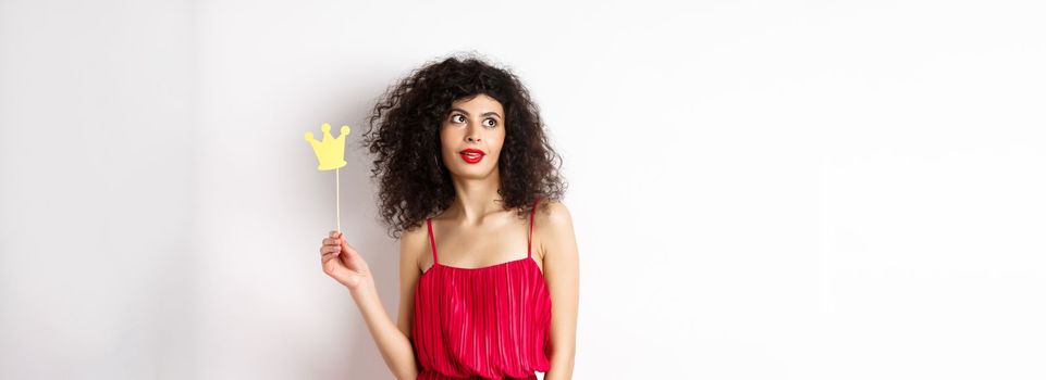 Stylish woman with curly hair in red dress, holding queen crown on stick and looking aside, standing on white background.