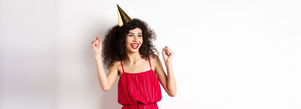 Happy birthday girl celebrating, wearing party hat and red dress, dancing and having fun, standing against white background.