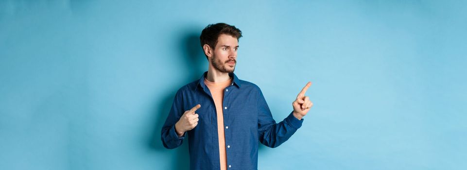 Image of young caucasian man looking and pointing aside, showing advertisement, standing on blue background.