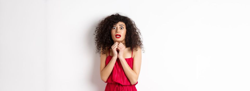 Scared caucasian woman trembling from fear, wearing red dress and staring anxious at camera, standing over white background.