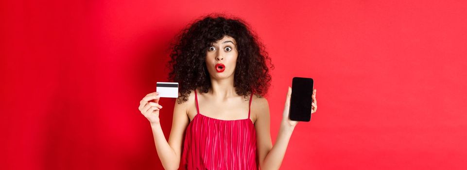 Online shopping concept. Beautiful woman in red dress and lipstick, showing empty phone screen and plastic credit card, standing on studio background.