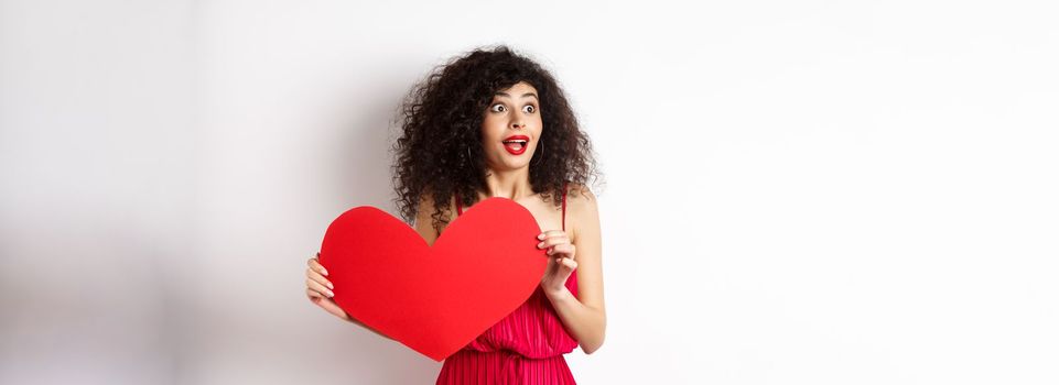 Valentines day. Young woman looking surprised left, seeing something amazing on lovers day, found true love, holding big red heart on chest, white background.