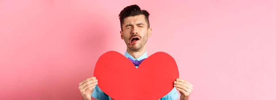 Valentines day concept. Sad and lonely man feeling heartbroken, being rejected, showing big red heart cutout and crying from break-up, standing on pink background.