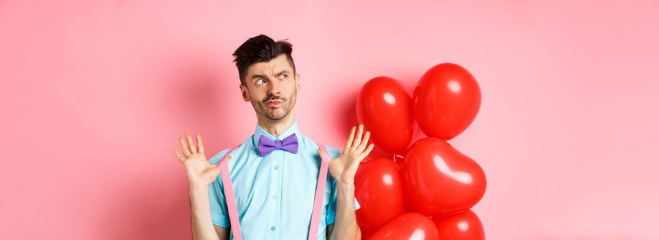 Valentines day concept. Pensive young man in bow-tie, raising hands up and looking left while thinking, making decision, standing on romantic pink background and heart balloons.