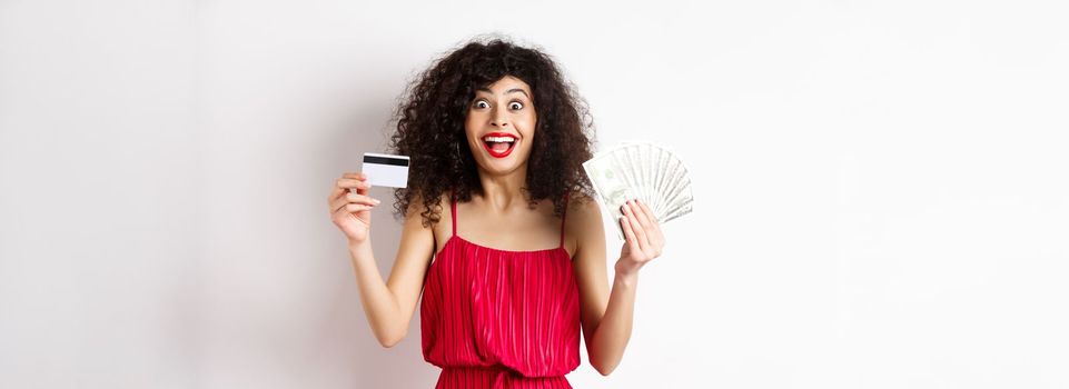 Happy attractive woman in red dress, screaming of joy and showing plastic credit card with money, winning prize, standing over white background.