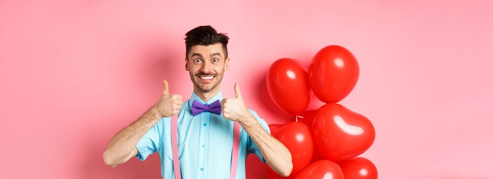 Valentines day concept. Smiling caucasian man showing thumbs up in approval, recommending special deal for lover, standing near cute red hearts balloons and pink background.