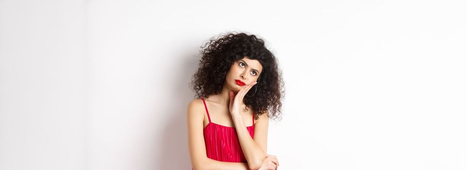 Annoyed and bored young woman with curly hair, look away distressed, lean face on hand, standing bothered in red dress on white background.