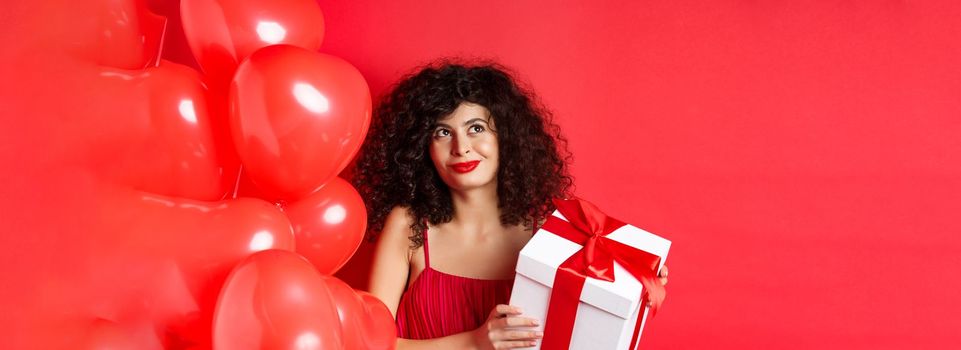 Valentines day and love concept. Beautiful woman with romantic makeup, holding gift box from secret admirer and looking at upper left corner dreamy, standing with heart balloons on red background.