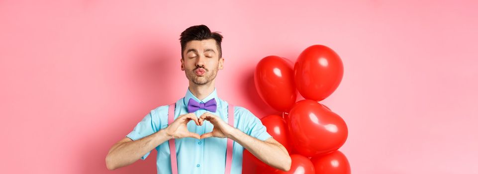 Valentines day concept. Passionate guy showing kissing face and heart gesture, waiting for love on romantic pink background.