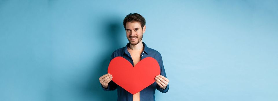 Handsome young man smiling, showing big red heart postcard for Valentines day, looking at camera happy, standing against blue background.