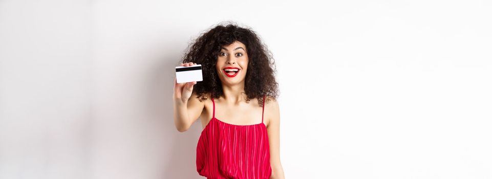 Shopping. Excited stylish lady with makeup, red dress, showing plastic credit card and smiling amazed, standing against white background.