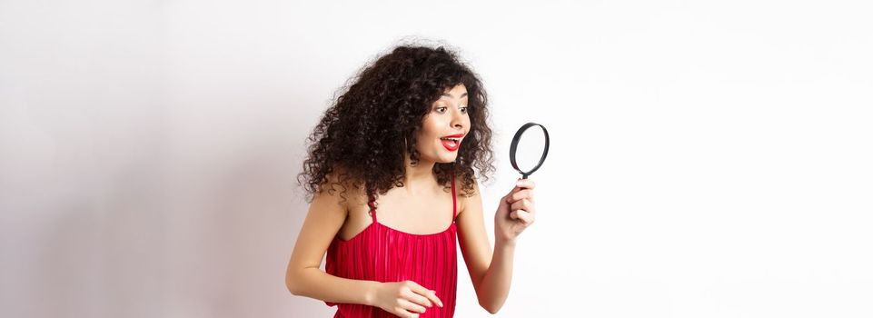 Beautiful woman in red dress and makeup looking at something with magnifying glass, checking out interesting promo, standing over white background.