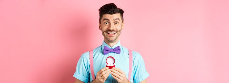 Valentines day. Romantic man in bow-tie showing engagement ring and smiling, asking to marry him, making proposal to lover on pink background.