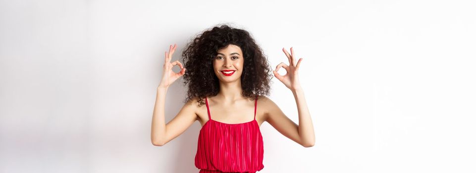 Beautiful woman in red dress, smiling and showing okay gestures, like something good, praising good job, standing over white background.