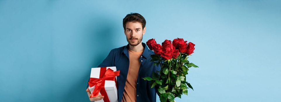 Romance and valentines day. Man presenting bouquet of red roses to lover. Boyfriend bring flowers and gift on romantic date, standing over blue background.