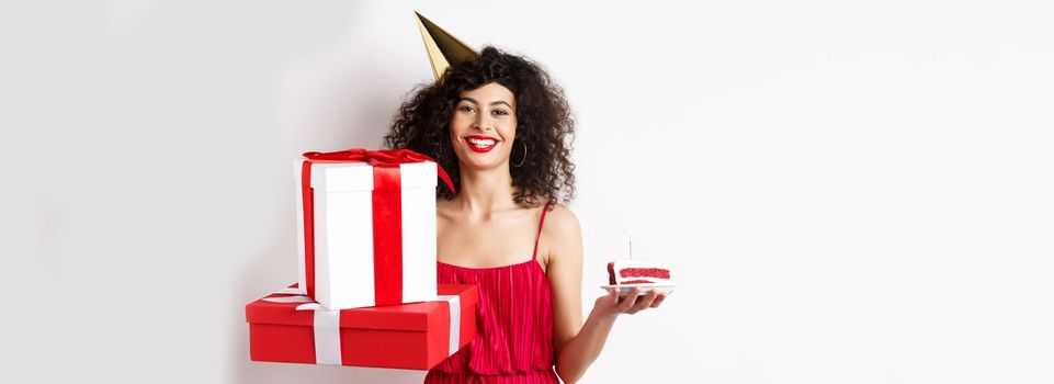 Happy birthday girl in red dress, celebrating and holding gifts with bday cake, standing on white background.