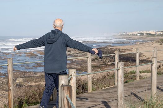 Grandfather on morning walk on a wooden path in the coast exercising and trying to balance with arms open.