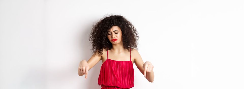 Sad and disappointed curly-haired woman in red dress, frowning and looking down jealous, pointing at promo, standing over white background.