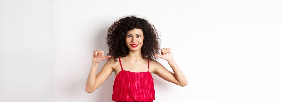 Confident young woman in elegant red dress, pointing at herself and smiling, self-promoting, standing over white background.