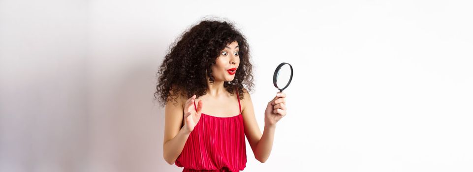 Excited woman in red dress look through magnifying glass and smiling, found interesting promo, standing on white background.