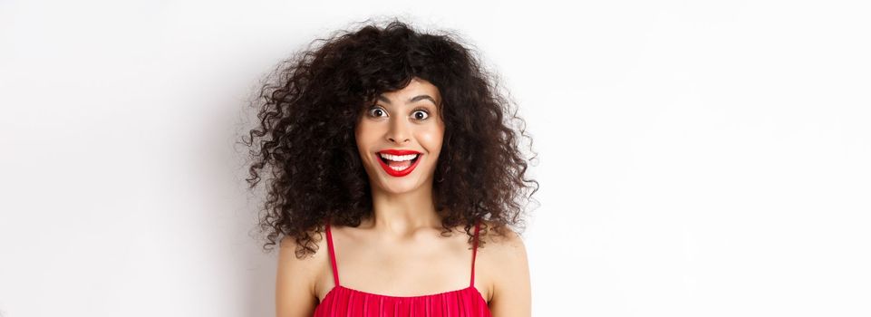 Close-up of happy lady with curly hair and red lips, raising eyebrows and looking surprised at camera, standing over white background.