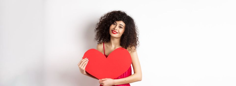 Romantic tender woman with curly hair, hugging big red heart and smiling, look with love, standing against white background.