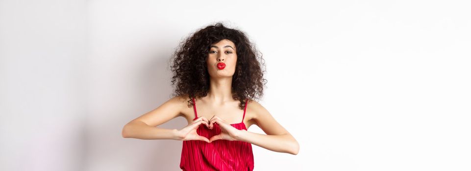 Beautiful lady with curly hair, red dress, showing heart symbol and pucker lips for kiss, love you gesture, standing on white background.