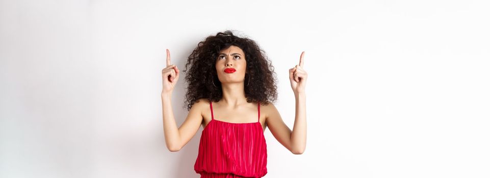 Angry and grumpy woman with curly hair, wearing red dress, frowning and looking up disappointed, standing over white background.