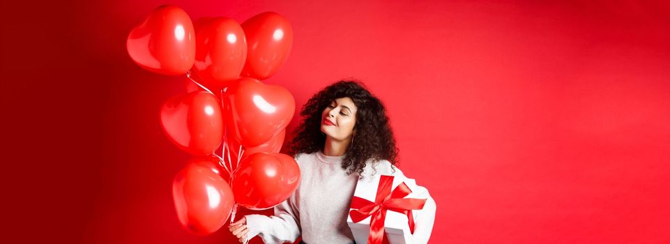 Romantic woman looking dreamy at heart balloons from lover, holding Valentines day gift in cute wrapped box, standing on red background.