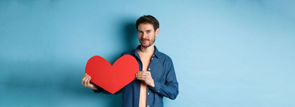 Handsome bearded guy showing valentines day cutout and smiling, standing in casual clothes over blue background. Copy space