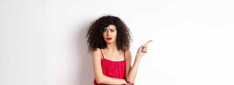 Arrogant young woman with curly hair, wearing red dress, frowning and complaining, pointing finger left at promo, standing over white background.