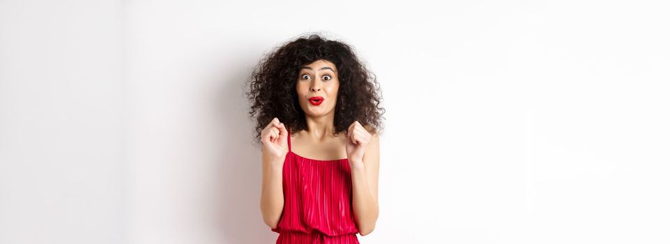 Excited smiling woman eager to try something, jumping from amusement and smiling, wearing red dress, standing on white background.