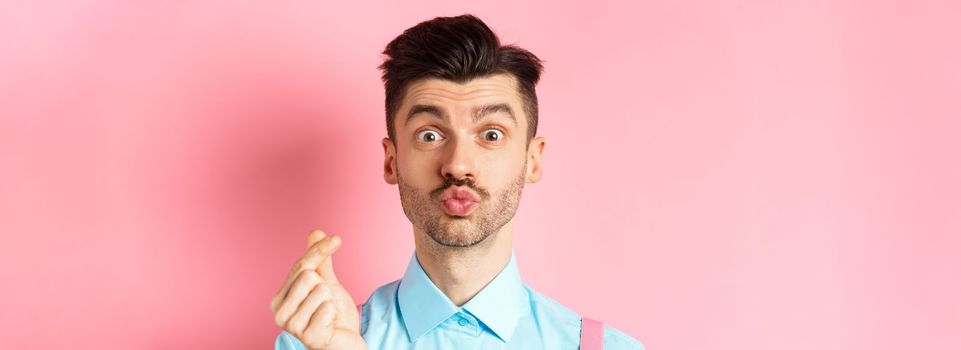 Valentines day concept. Silly guy waiting for kiss and showing finger heart gesture, express love and sympathy, pink background.