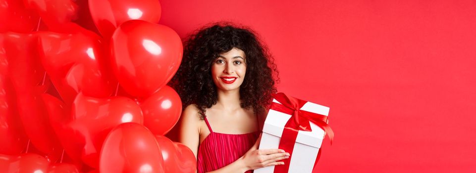 Holidays and celebration. Beautiful woman with curly hair, standing near heart balloons, holding gift box and smiling happy, white background.