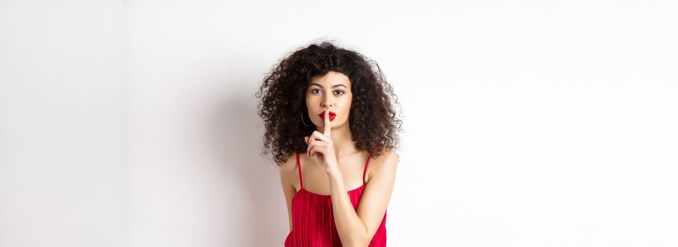 Beautiful woman with curly hair, wearing red dress, hushing at camera, telling secret, shushing at you, standing over white background.