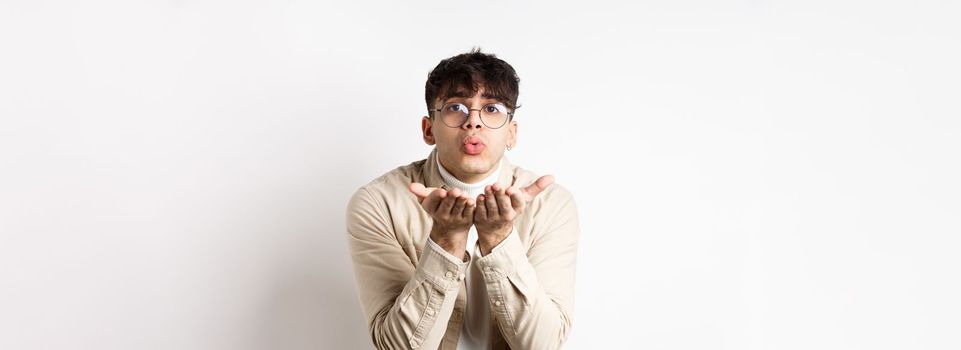 Cute boyfriend sending air kiss at camera on happy Valentines day, looking tender at lover, standing in casual clothes and glasses on white background.