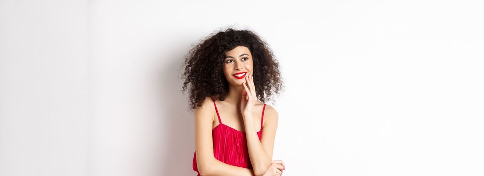 Dreamy elegant girl with curly hair, wearing red dress and makeup, looking left and smiling at logo, standing on white background.