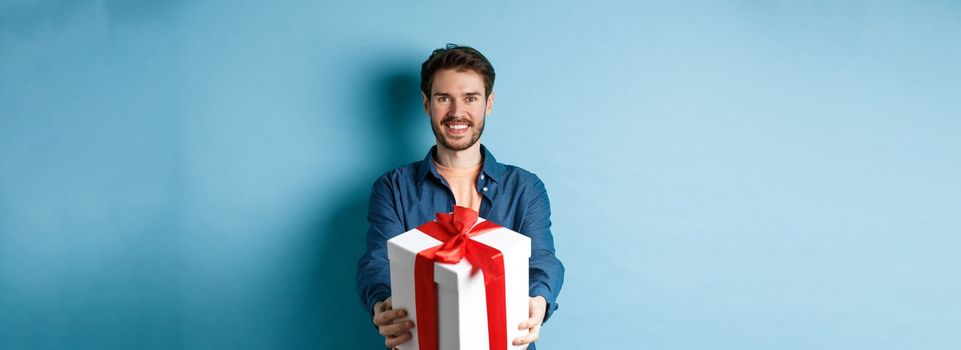 Valentines day. Handsome smiling man extending hands with gift box, wishing happy holiday. Guy making surprise present and looking cheerful, standing over blue background.