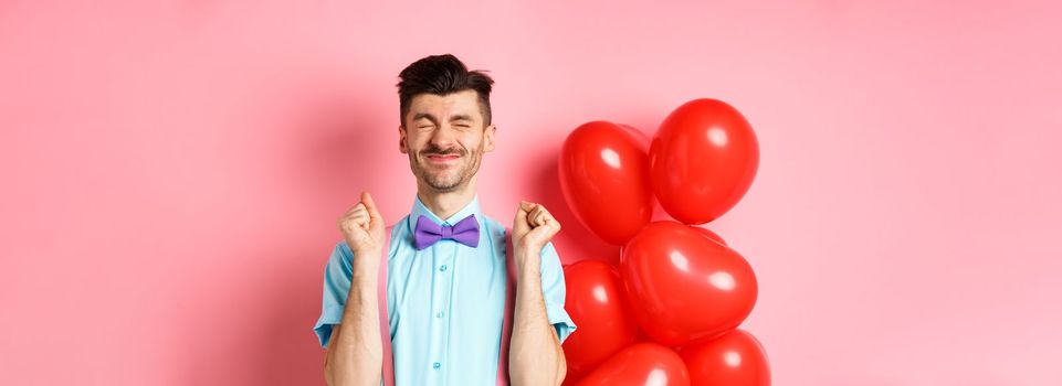 Valentines day concept. Cheerful guy jumping from excitement before romantic date, standing near big red hearts balloons and celebrating, pink background.