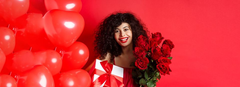 Happy Valentines day. Romantic girl with presents from lover, holding bouquet of roses and gift box, standing near cute red hearts on studio background.